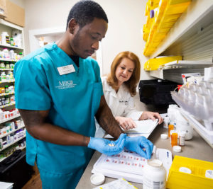 A pharmacy student prepares medication as a pharmacist oversees and reviews paperwork in a pharmacy workspace.
