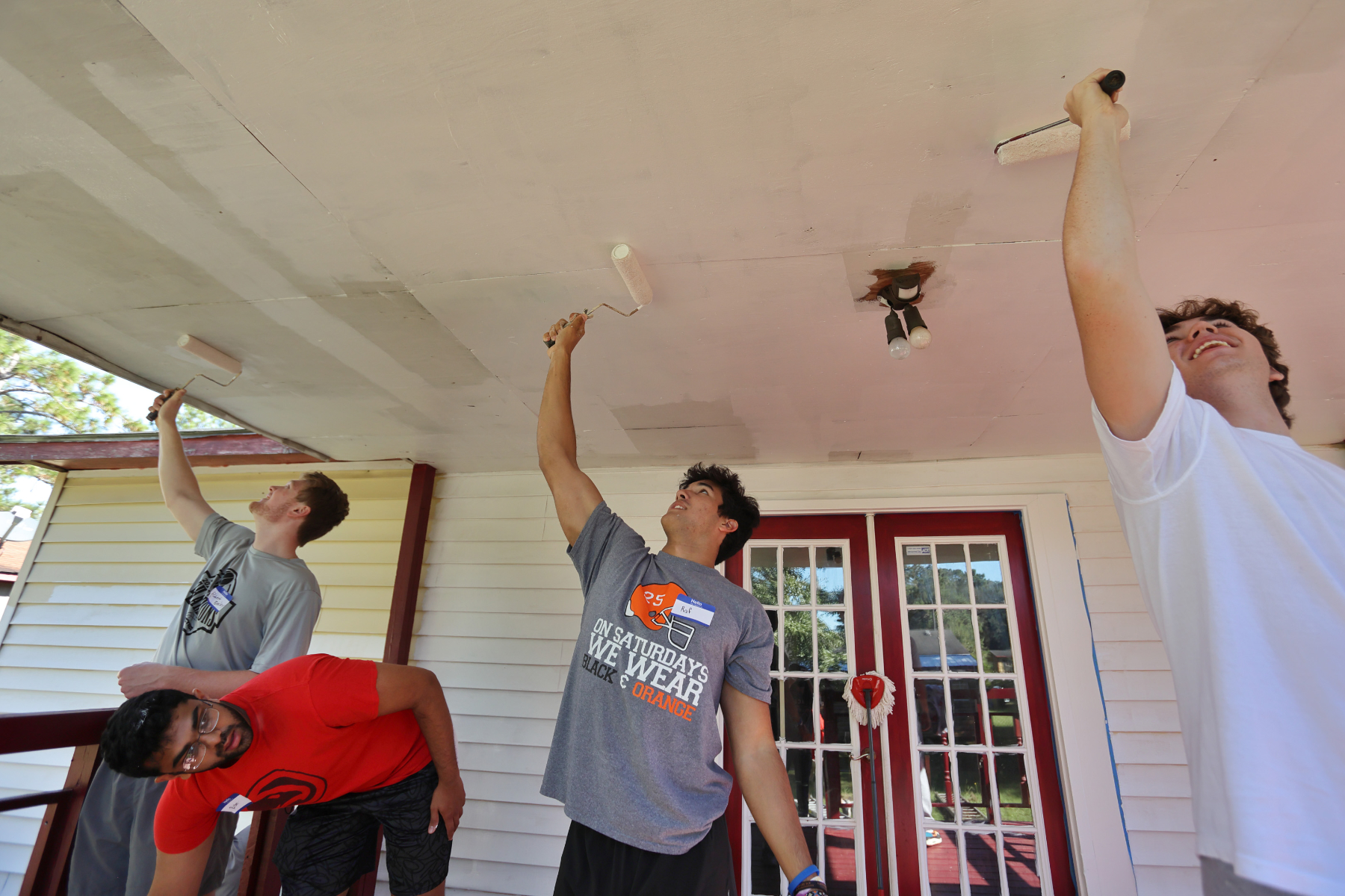 Three Mercer students reach overhead painting the ceiling of a home's porch. Another student is leaning down as if to grab something.