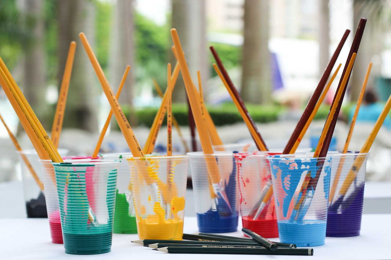 eight clear plastic cups holding different colored paint. paint brushes stick out of the top of the cups.