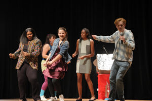 four women dance on stage while a man sings into a microphone
