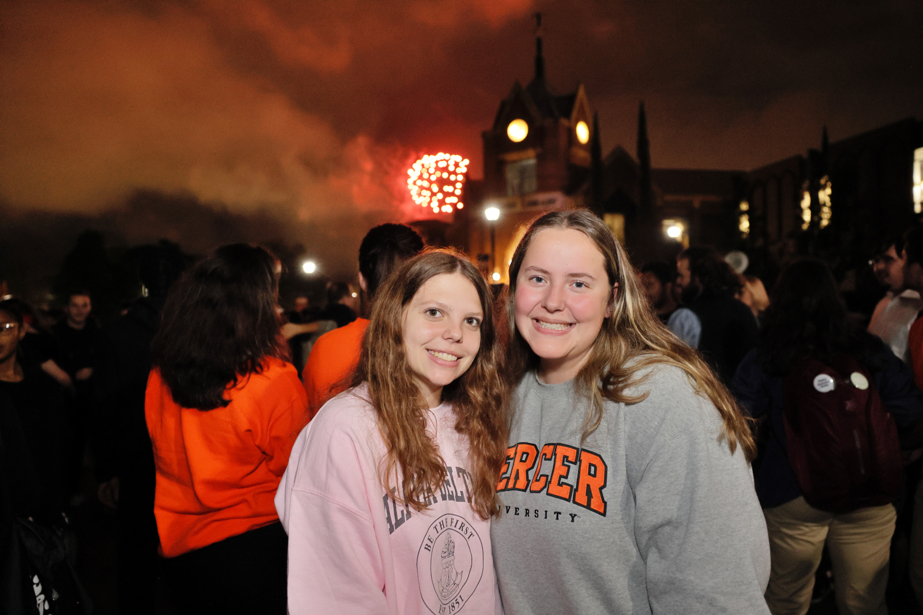 two college students smile while fireworks go off behind them