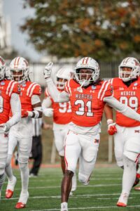A group of Mercer University football players in orange and white uniforms running on the field during a game. One player with number 31 prominently visible is leading and gesturing with enthusiasm.