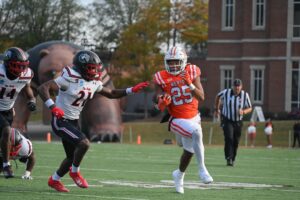 A football player wearing an orange and white Mercer uniform with the number 25 on it is running with the ball. Two defensive players in white, black, and red uniforms from "The Webb" are attempting to tackle him. The game takes place on a college campus, with a large inflatable bear in the background and an official running behind the players.