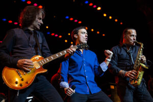 man sings on stage between a man playing guitar and a man playing saxophone