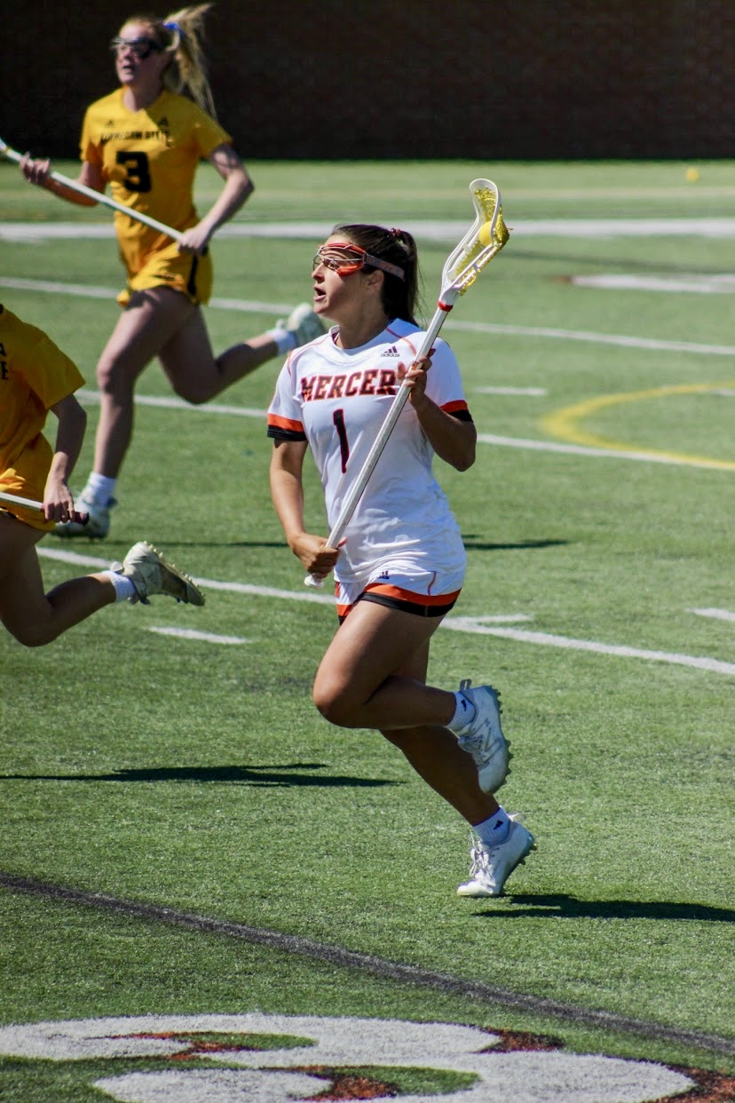 A women's lacrosse player in a white and orange uniform labeled "MERCER" is running on a field, holding a lacrosse stick and looking upwards, with another player in the background.