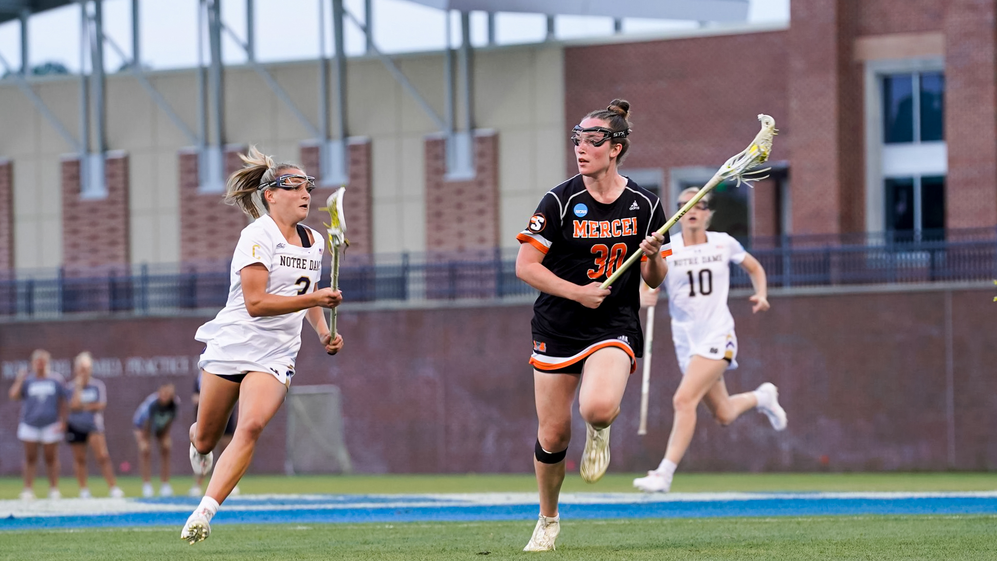 Two women's lacrosse players in action on a field; one player in a white and light blue uniform chases another in a black and orange uniform, who is carrying the ball with a lacrosse stick. A sports complex is visible in the background.