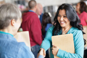 A person with long black hair, wearing a turquoise shirt, and smiling broadly, shakes hands with another person holding a clipboard, whose back is to the camera. They are in a busy indoor setting with several other people in the background.