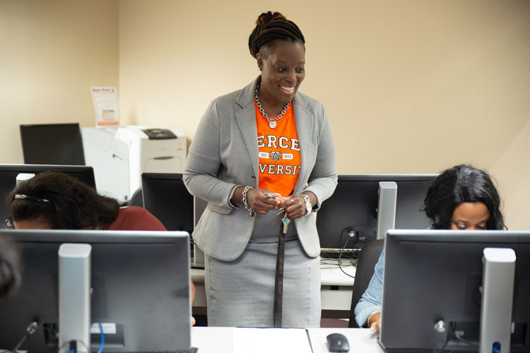 A person wearing a necklace and an orange Mercer University T-shirt under a gray suit jacket smiles while standing in an office-like classroom setting with multiple students working on desktop computers.