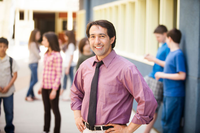A teacher smiling at the camera with students in the background in a school hallway.