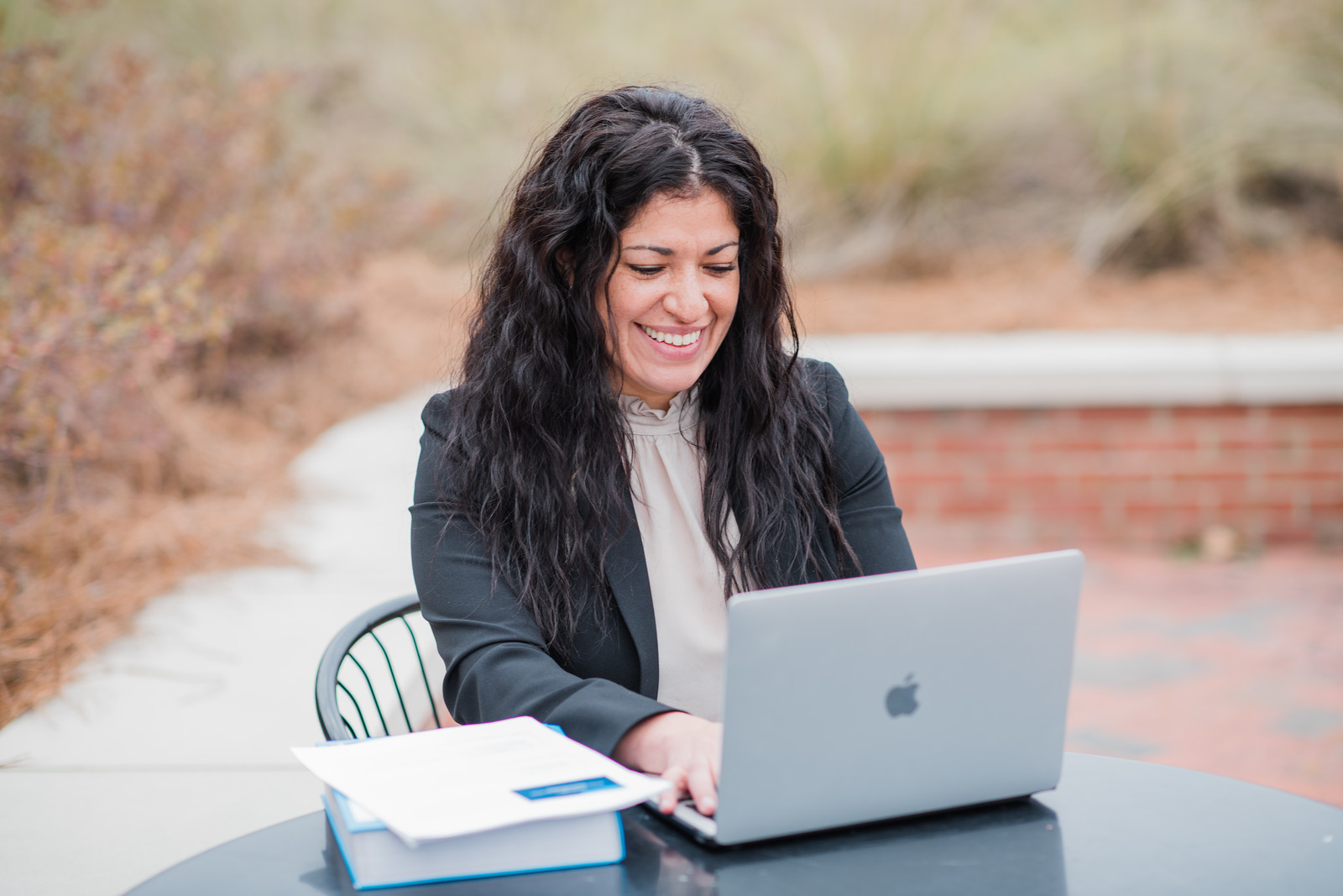 Person sitting at a metal table outdoors, working on a laptop and smiling while looking at the screen. A textbook with a piece of paper on top is also on the table. The background shows soft focus greenery and a sidewalk.