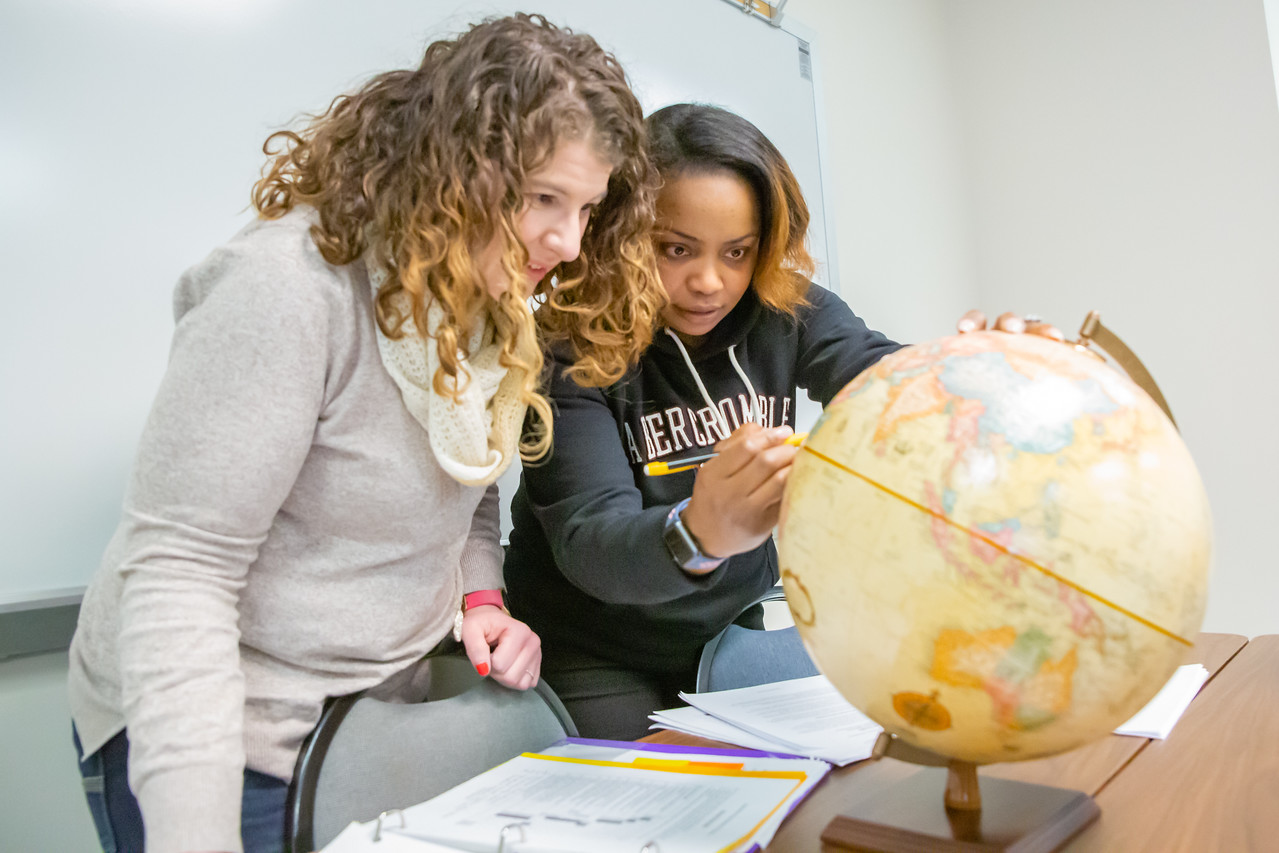 Two students are examining a globe closely in a classroom setting, with books and papers spread out on the table in front of them, indicating a study or learning session.