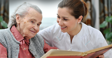 A caregiver, wearing a white uniform, is sitting beside an elderly person, who is wearing a gray cardigan. They are both looking at a book that the caregiver is holding. The setting appears to be a cozy room.
