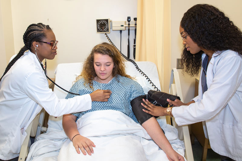 Two Mercer students wearing white lab coats examine a patient in a hospital bed; one listens to the patient's heart with a stethoscope while the other checks the patient's blood pressure.