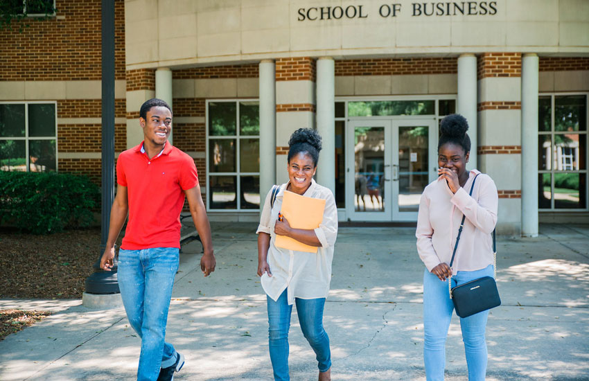 Three students walking and laughing together outside the "School of Business" building on a sunny day.