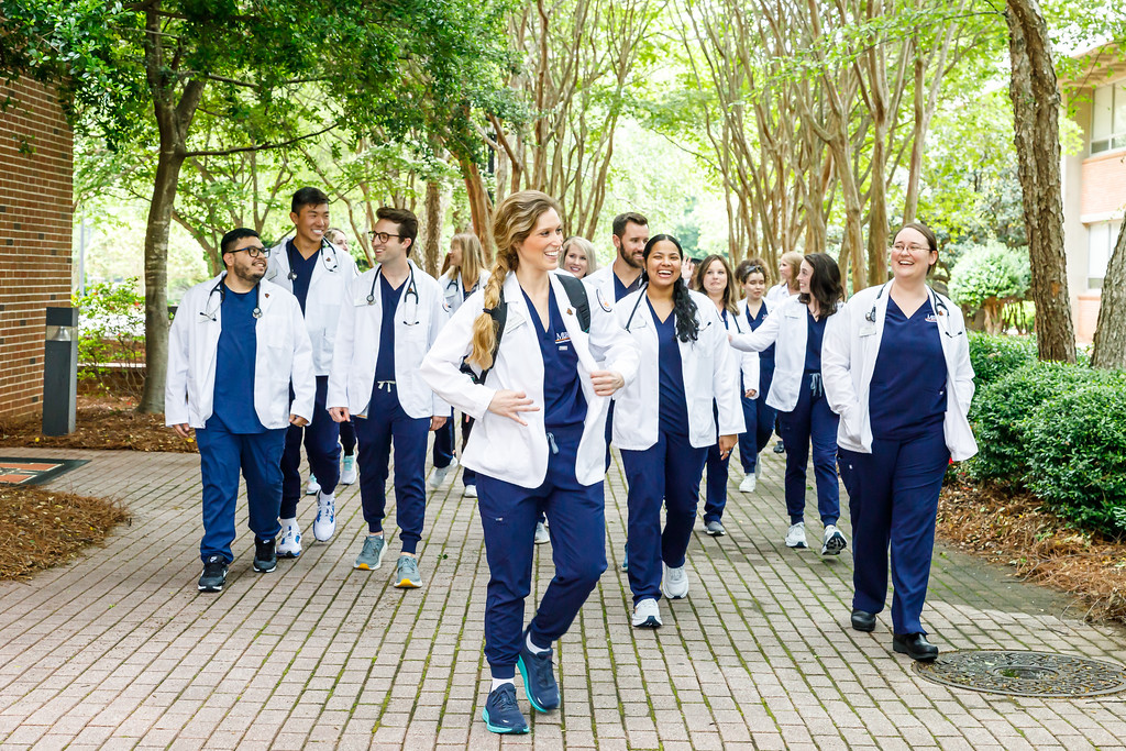 A group of individuals in white lab coats and dark blue scrubs walking and talking together along a tree-lined pathway. Some are smiling and appear engaged in conversation. The environment suggests a university setting.