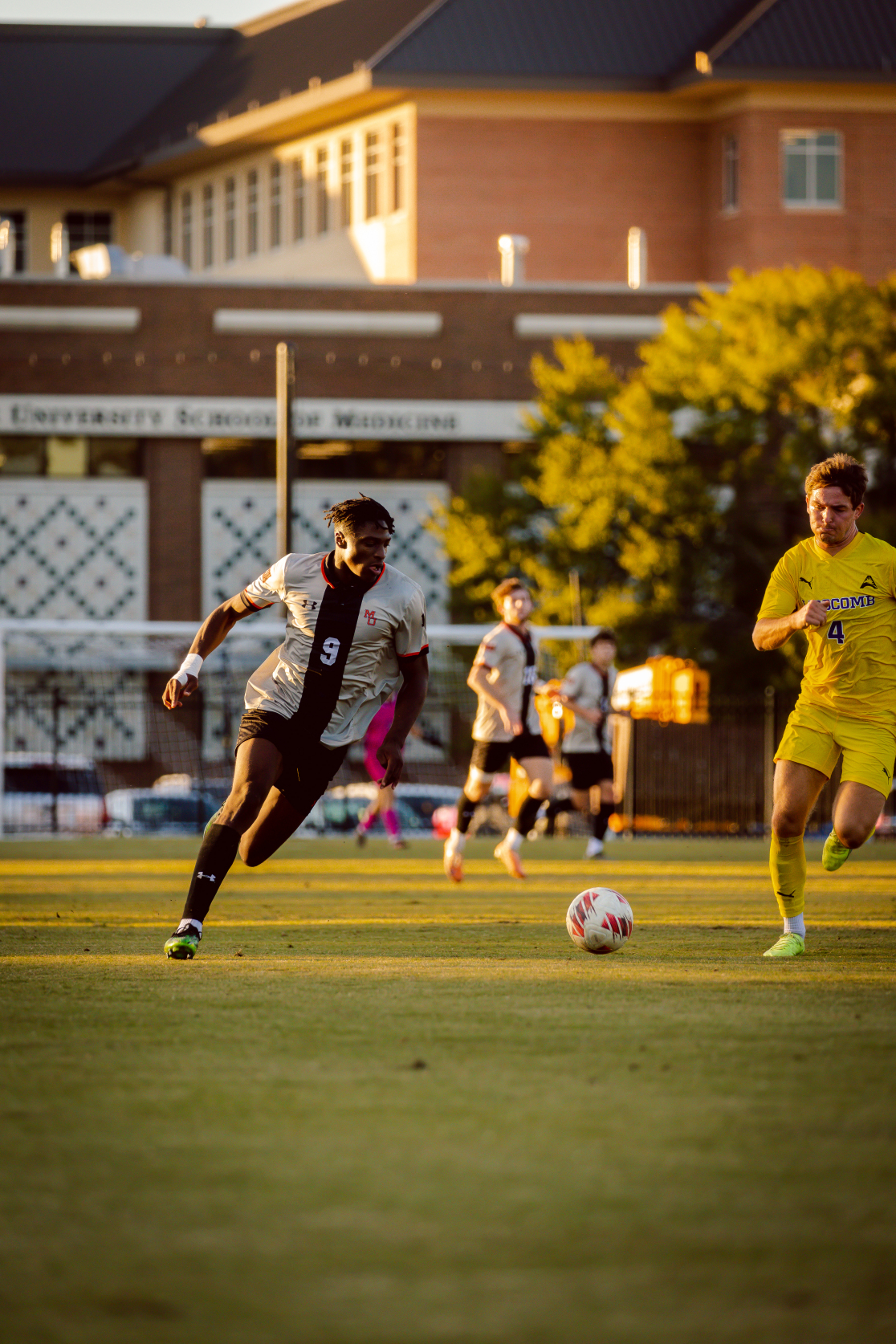 Mercer player number 9 runs to the left of the soccer ball during a soccer match. A player from the team in yellow, number 14, runs to the right of the ball. They are on a field flanked by a brick building under a setting sun.