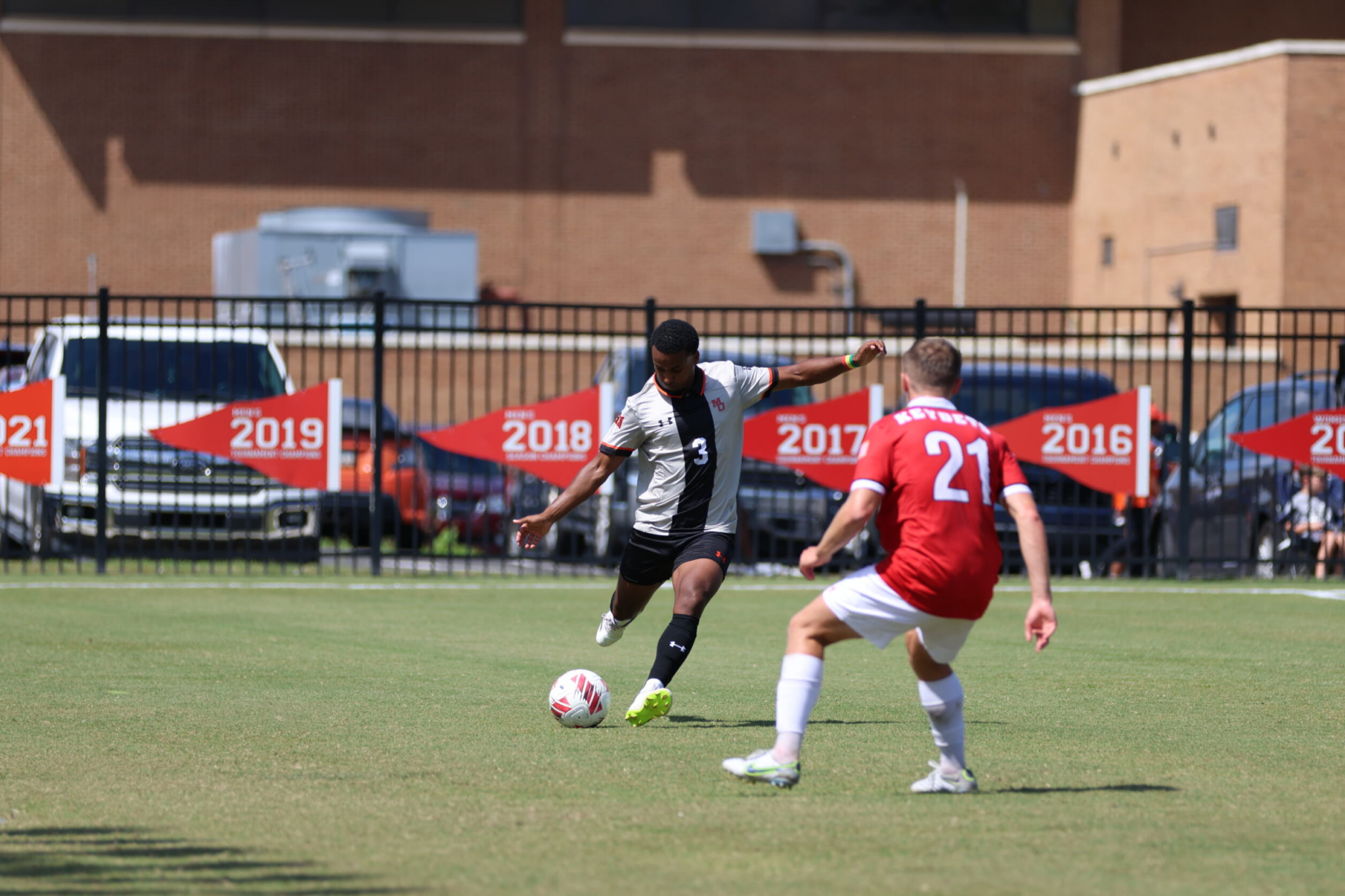 Two soccer players in action on the field; one in a black kit with the number 3 and the other in a red kit with the number 21, engaging in a duel for possession of a soccer ball.