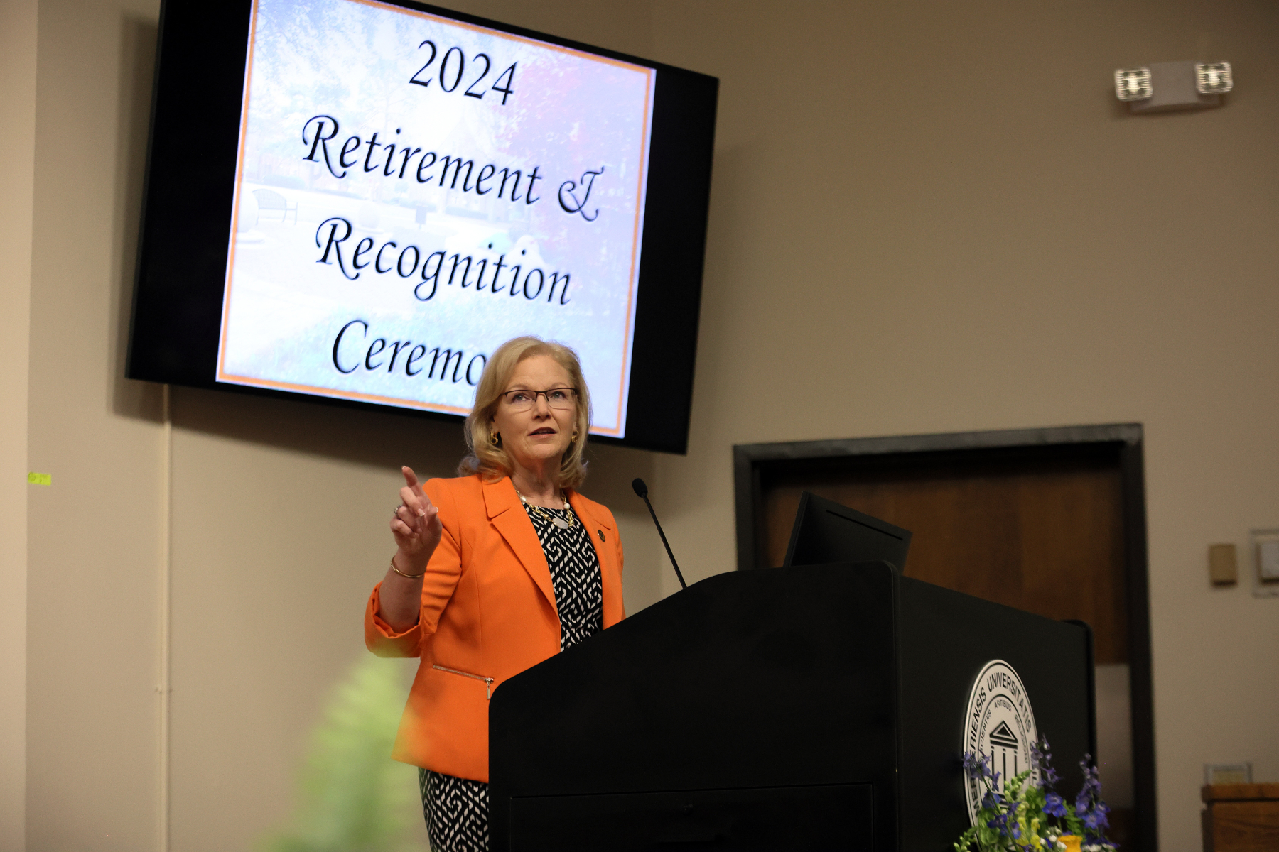 a woman wearing an orange blazer standing behind a podium talking and gesturing. The words "2024 Retirement and Recognition Ceremony" are shown a TV screen behind her.