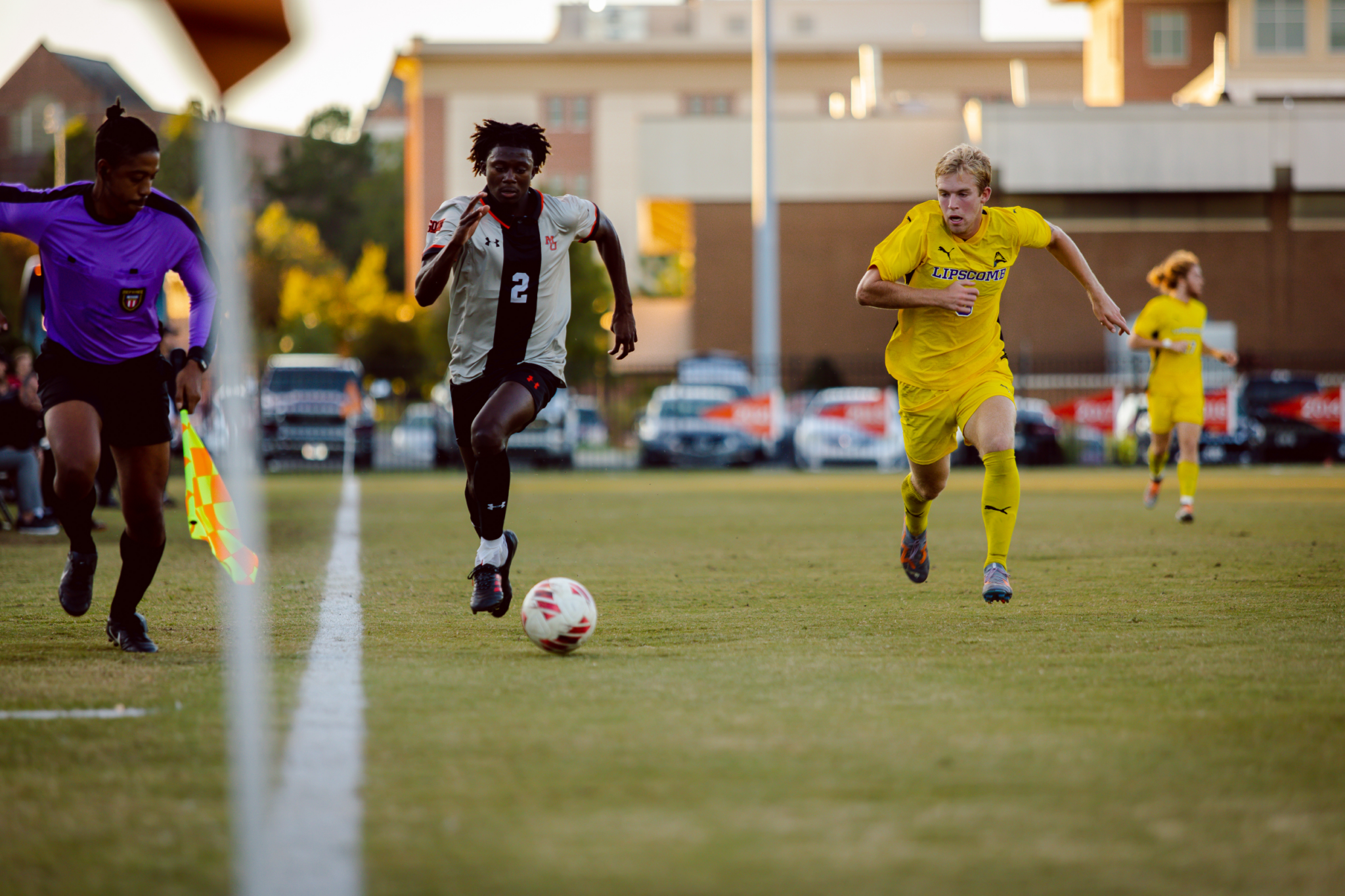 Soccer players in action on a grassy field during a game at sunset. A Mercer player is dribbling the soccer ball, closely followed by a player in a yellow uniform. A referee wearing purple is watching closely from the sideline.