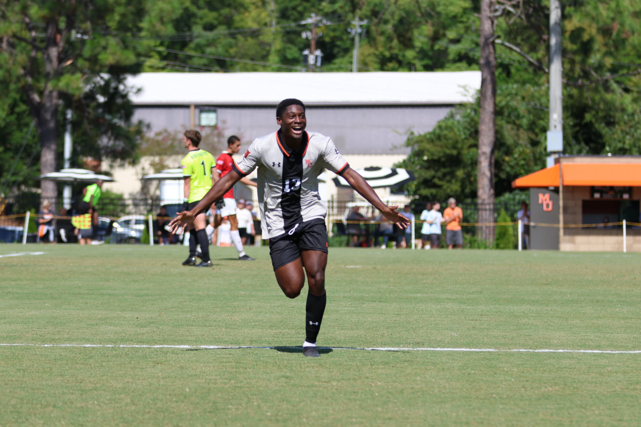 A soccer player in a black and gray Mercer uniform joyfully running on the field during a match, with other players and spectators in the background.