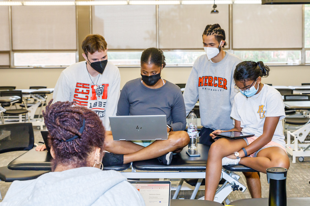 Four students in a classroom, two standing and two seated, all focused on a laptop or iPad and wearing face masks. They are engaged in what appears to be a group study or project session. The environment is equipped with desks and chairs typical of an educational setting.