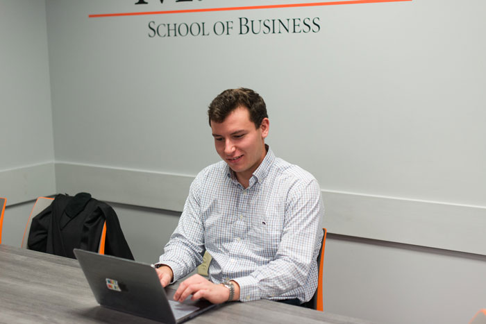 An individual sits at a table while working on a laptop. A backpack is in the next chair. On the wall, it says "School of Business"