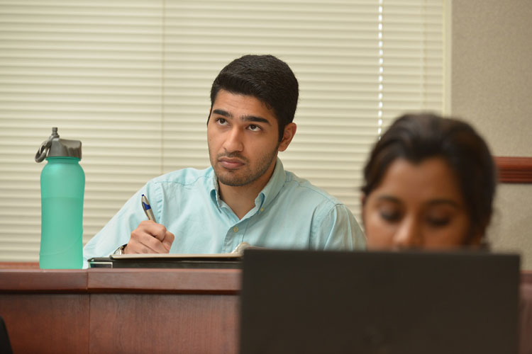 A student sitting at a desk looks up from taking notes. A student in front of him looks at a laptop