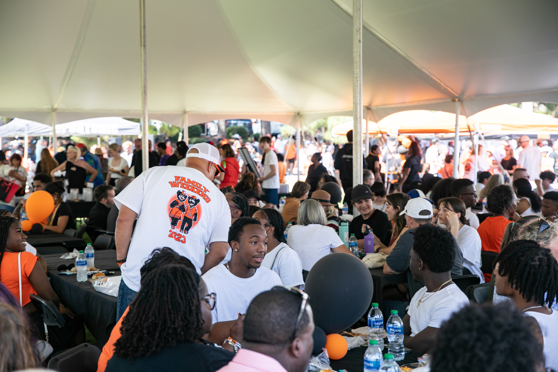 People sit at crowded tables under a large, white tent outside. One person is standing, and the back of his shirt features a picture of the Toby and Tot bear mascots and reads "Family Weekend 2023."