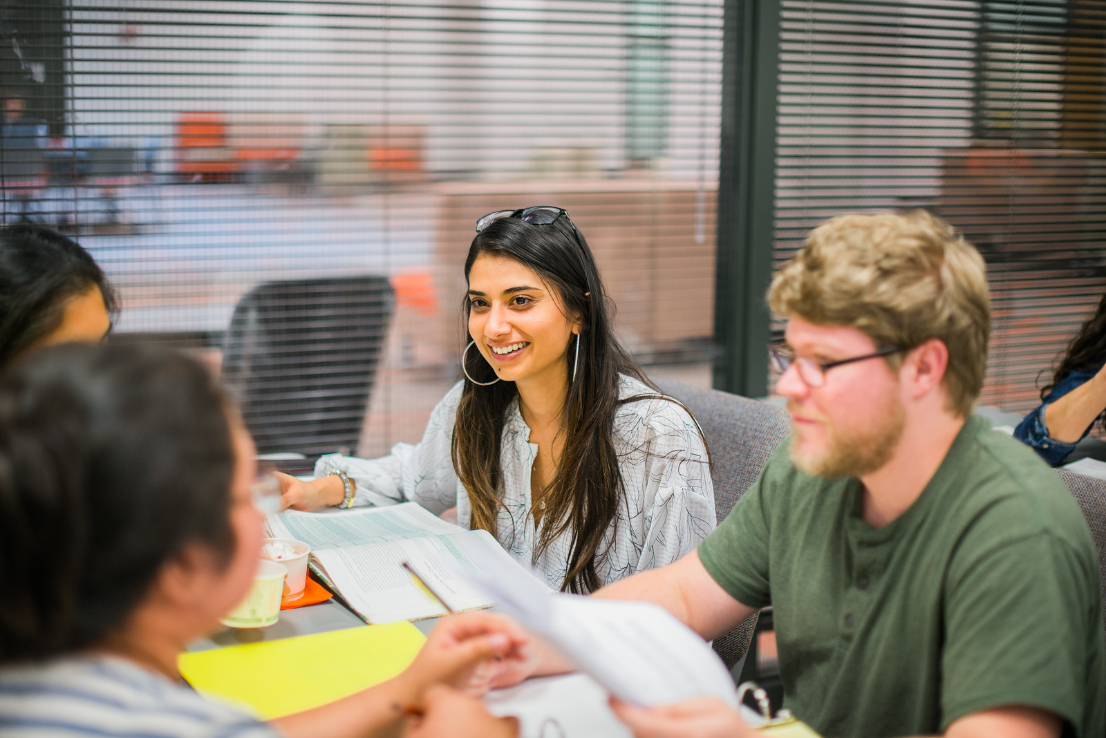 A group of students engaged in a study session around a table, smiling and discussing notes, in a bright room with a windowed background.