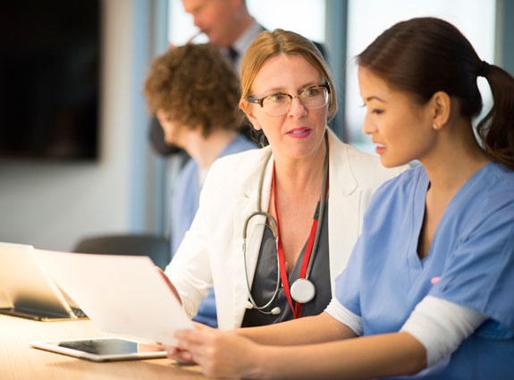 Two health care professionals, one wearing a lab coat with a stethoscope and the other in scrubs, are reviewing documents together at a desk in a medical office. There are additional colleagues in the background.