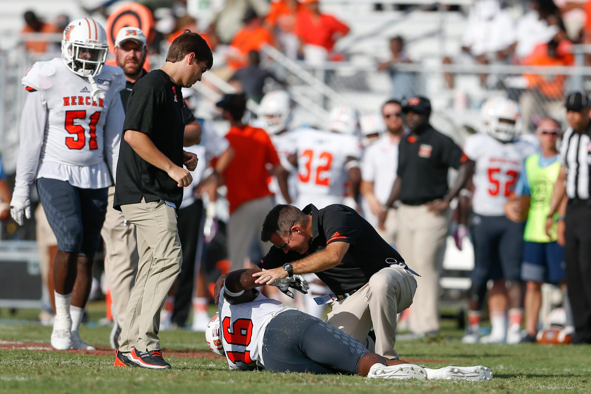 A Mercer football player wearing a jersey with the number "16" is laying on the ground, appearing injured, while being attended to by two individuals, likely team medical staff or coaches. Other players and staff members can be seen in the background. The scene suggests a pause in play for injury assessment.