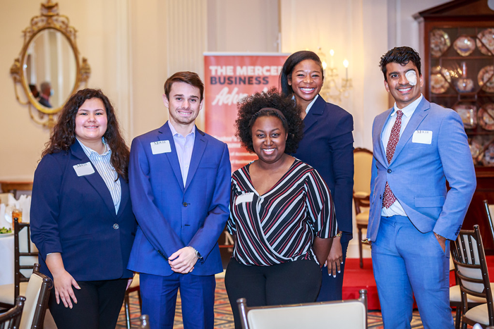 Five diverse individuals wearing business attire smile while standing in a large dining room. Behind them is a banner that says, "The Mercer Business Advantage."