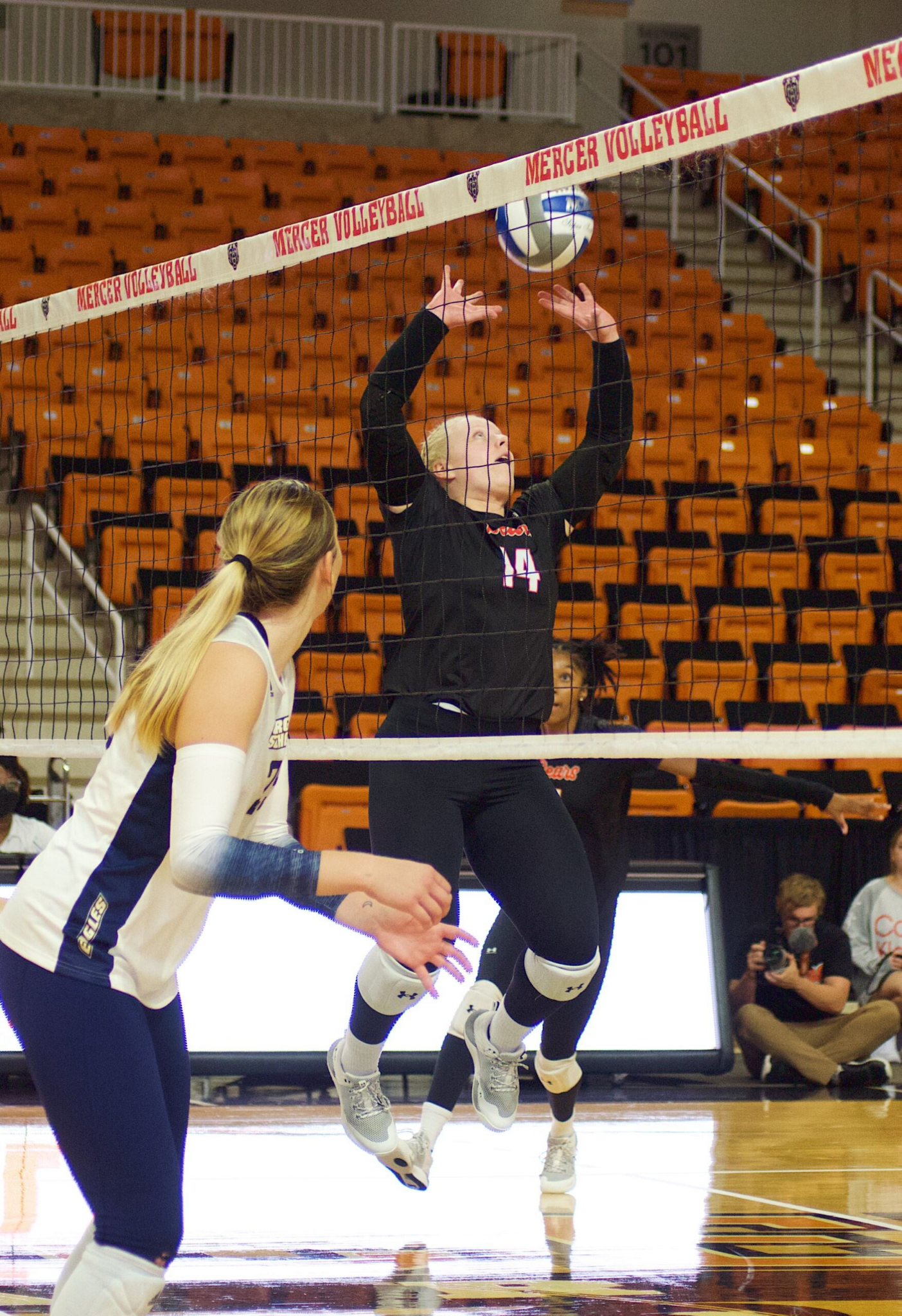 Two volleyball players in action during a game, with a Mercer player in a black uniform jumping to hit a volleyball over the net and another in a white and blue uniform preparing to return. They are in an indoor court with orange seats visible in the background.