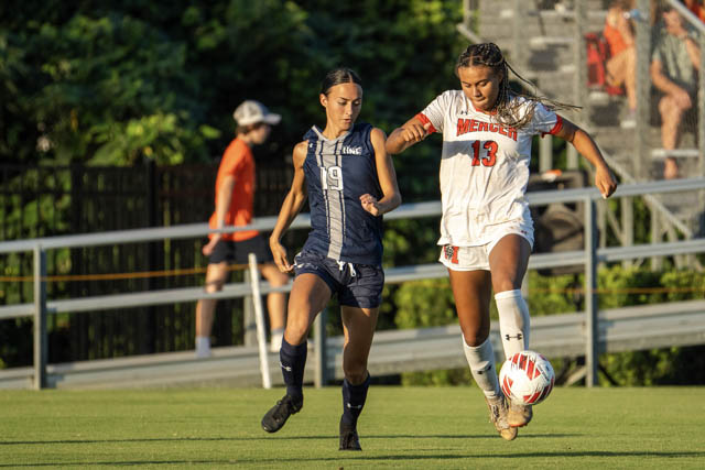 A Mercer women's soccer player dribbles a ball during a match. An opponent in a navy blue uniform closely follows her.