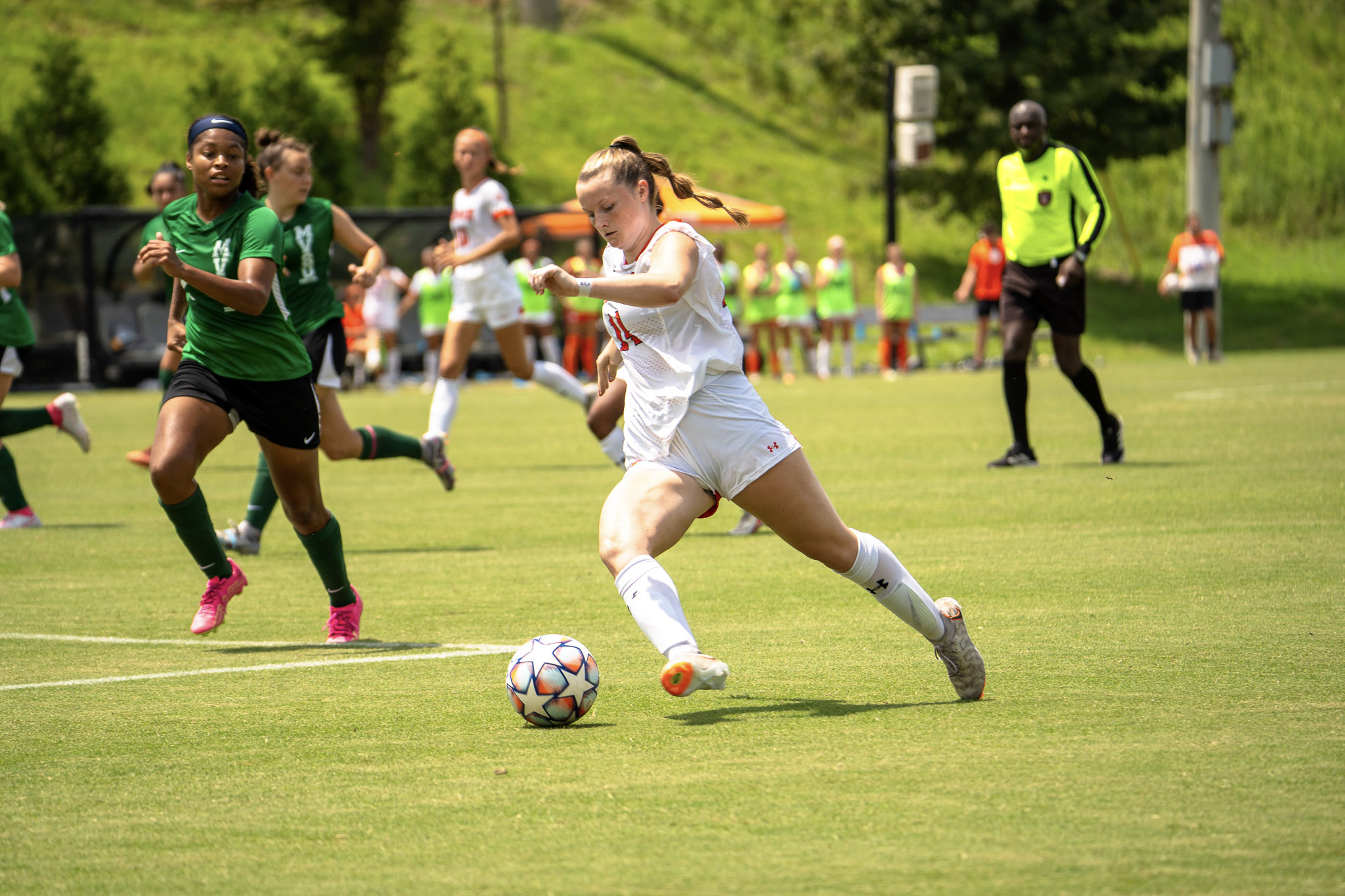 A Mercer women's soccer player goes to kick the ball during a match while an opponent wearing a dark green uniform approaches to challenge. A referee and spectators are visible in the background.