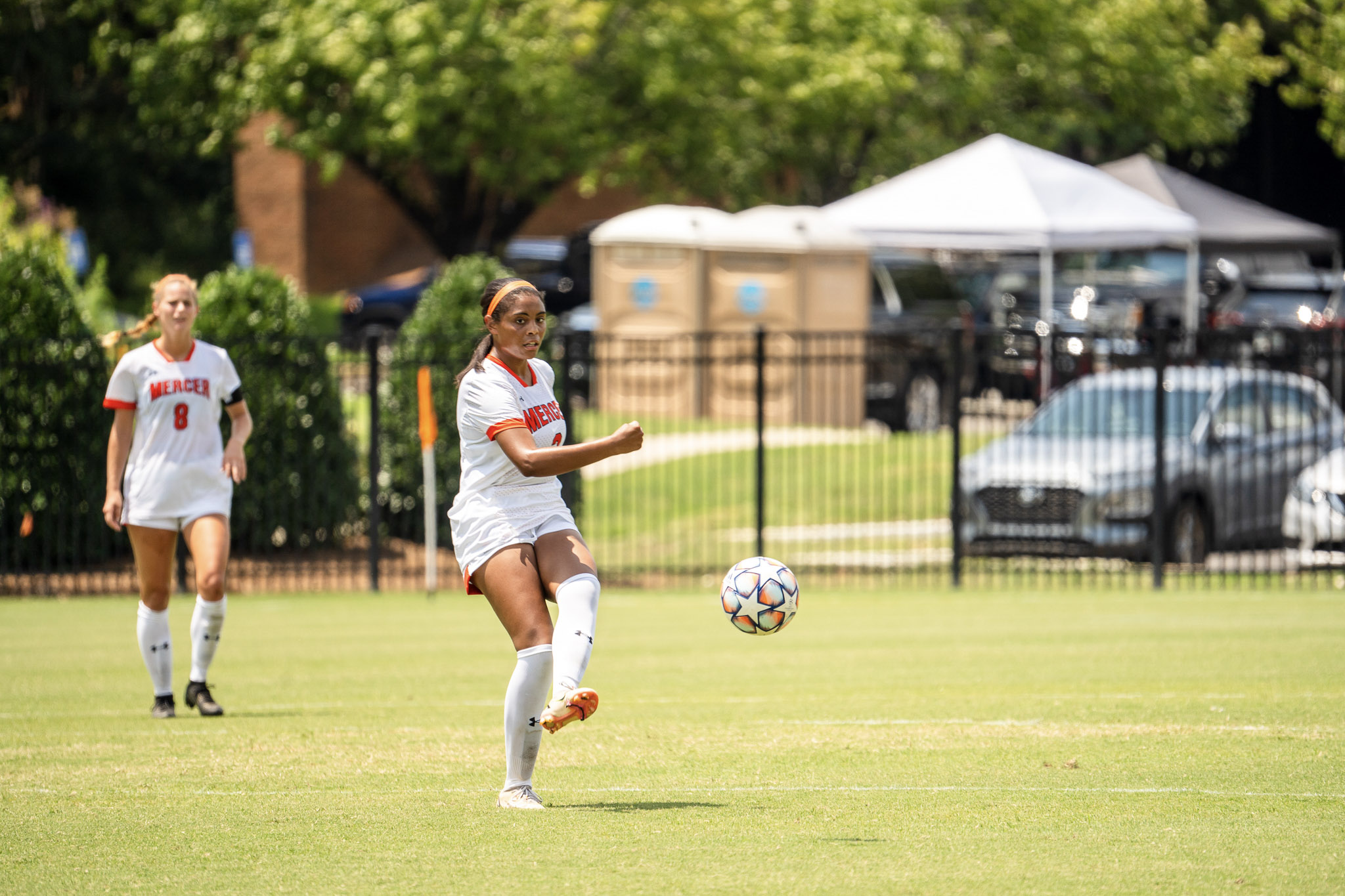 Two Mercer soccer players in action on a sunlit field. One player in the foreground is about to kick a soccer ball, while the other approaches from behind.