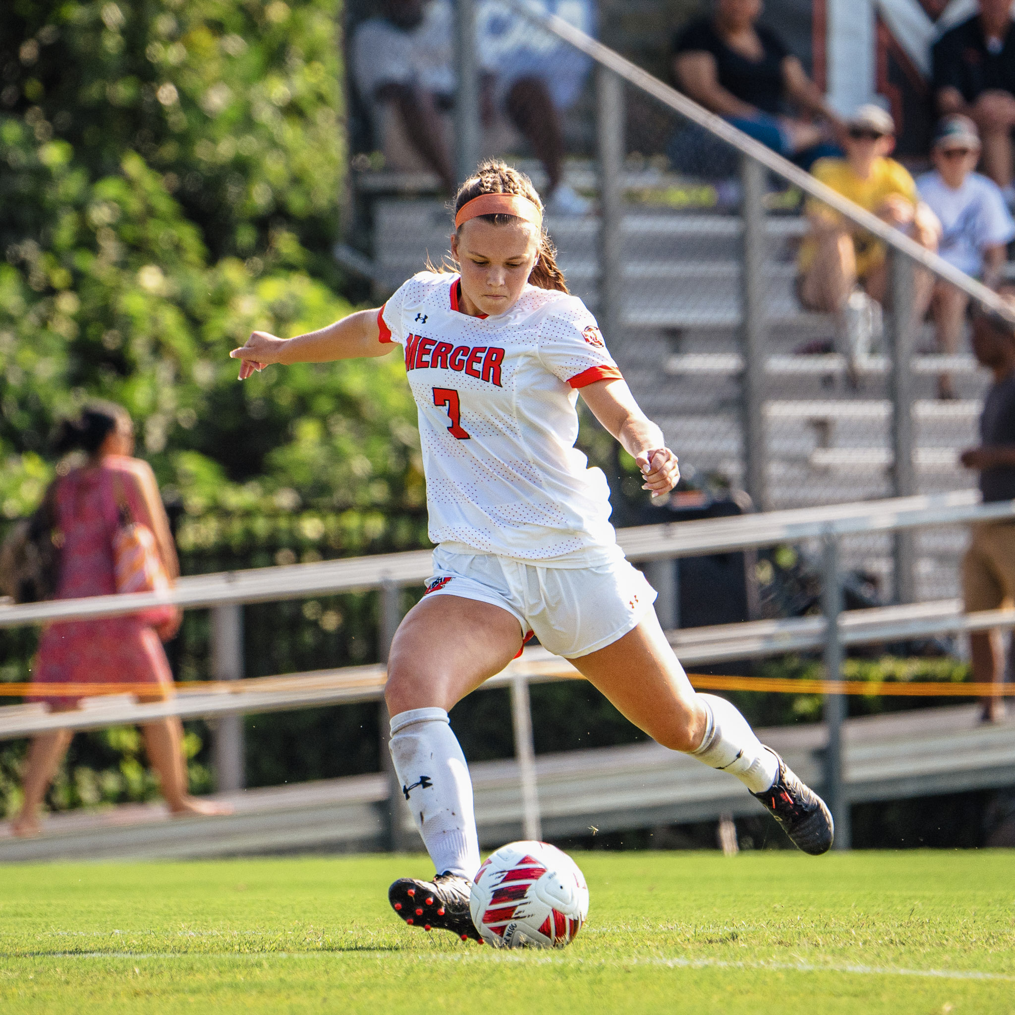 A Mercer women's soccer player dribbles the ball intently during a game, with spectators in the background.