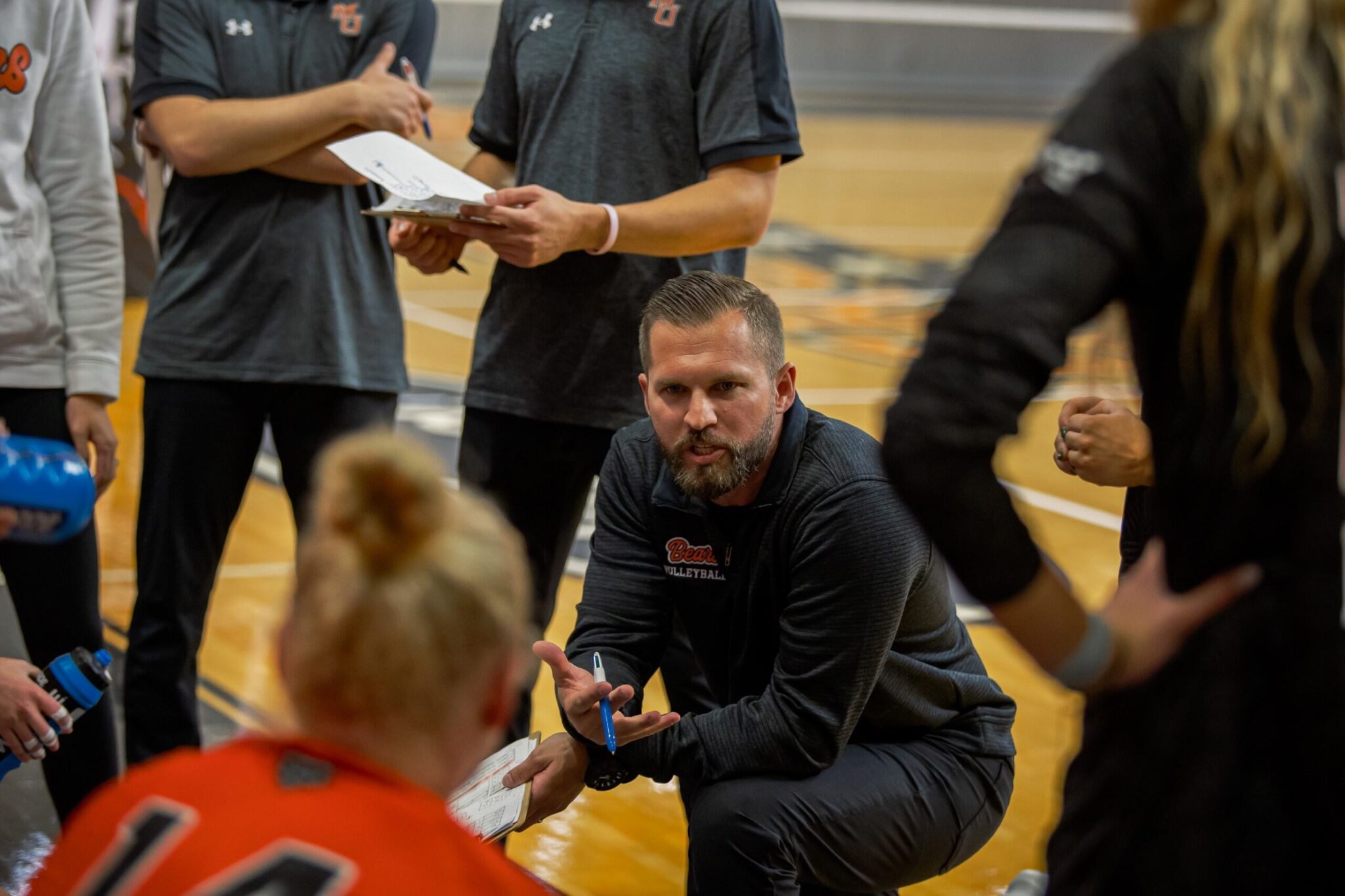 A Mercer coach is kneeling in a gymnasium, engaging with a group of basketball players seated around him. The coach is gesturing with his hand while holding a clipboard. The players are wearing orange jerseys.