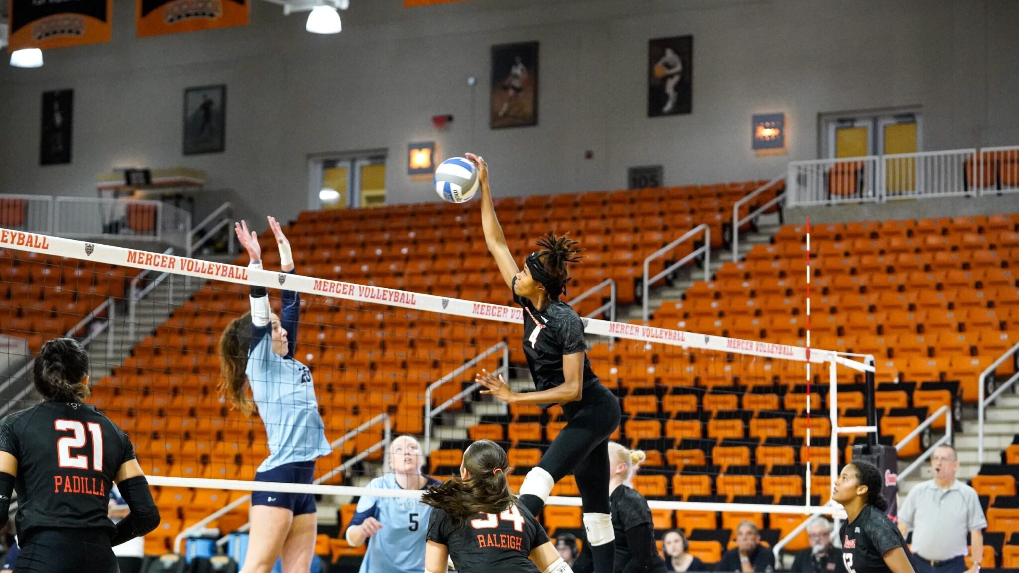 Two college volleyball teams in action on an indoor court, with one Mercer player in black and another opposing player in blue reaching up to hit a volleyball over the net. The court is surrounded by orange bleachers.