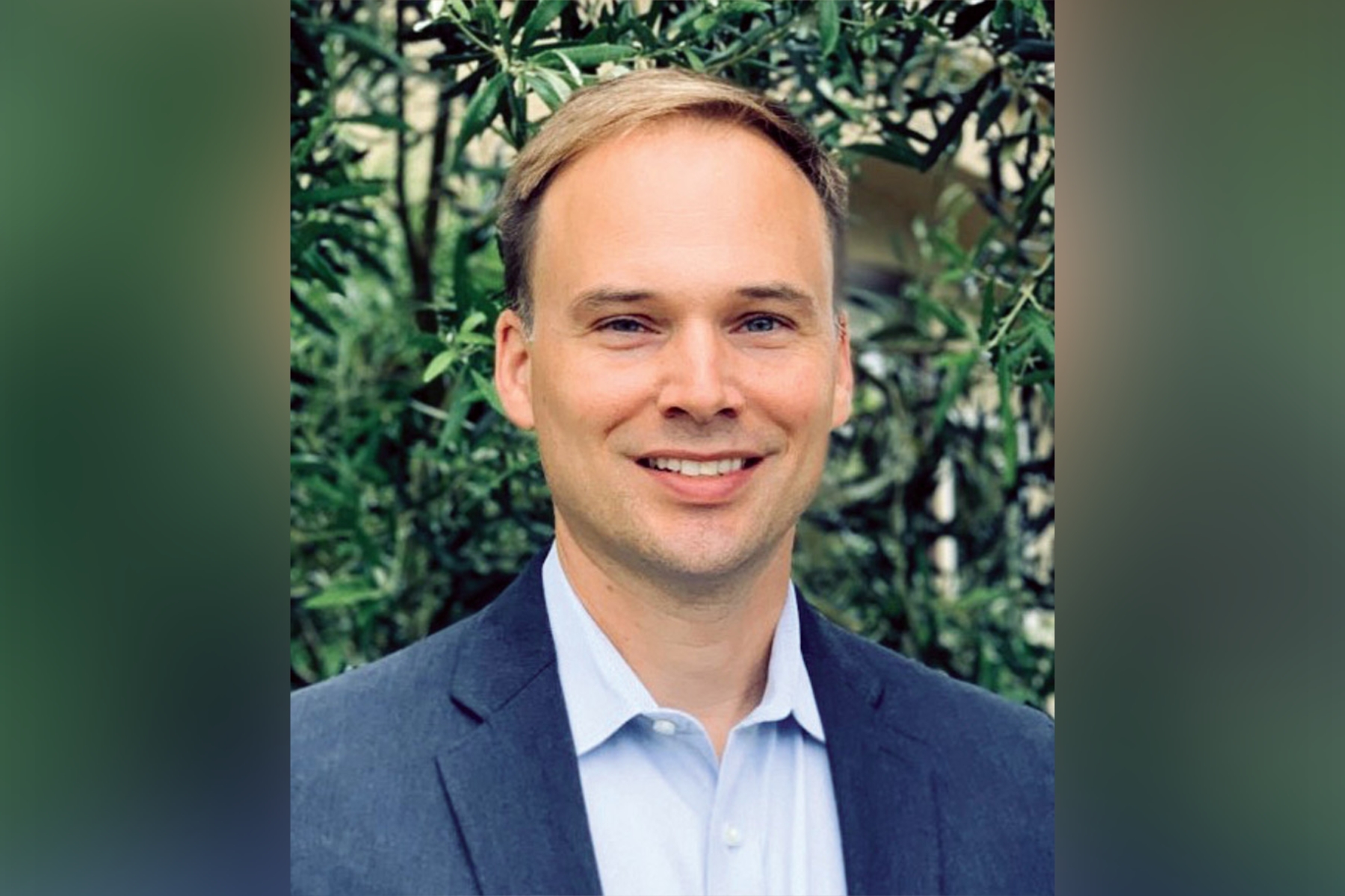 Headshot of John Compton wearing a blue suit and standing in front of trees.