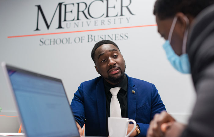 A man wearing a tie and blazer talks to a person seated to his right. They are in front of a wall that reads, "Mercer University School of Business."