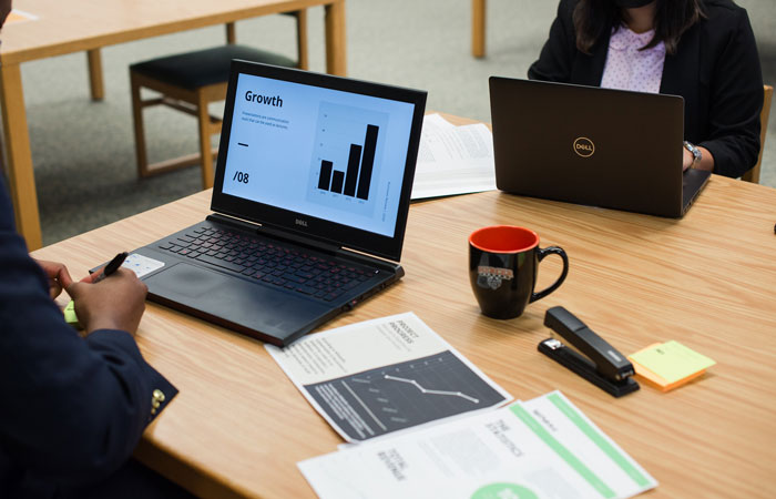 Two individuals, both partially visible, sit at a table. One person looks at data on a laptop screen, showcasing a chart titled "Growth" with rising bars. The other person works on a separate laptop. The table is covered with business documents, a black coffee mug with the Mercer logo, a stapler, and a yellow post-it note.