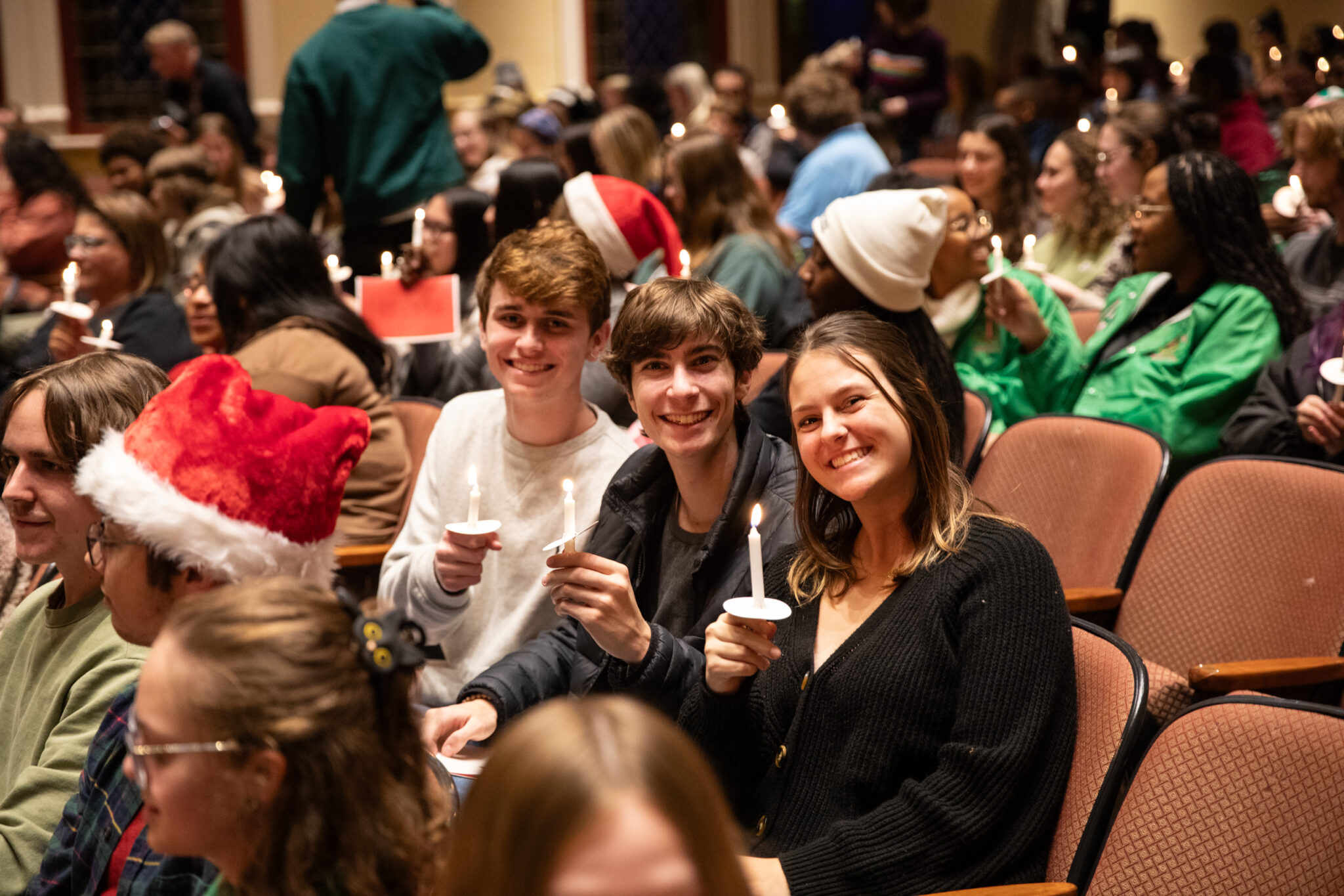 Three Mercer students hold candles while sitting in an auditorium filled with people. Some Santa hats are seen in the background.