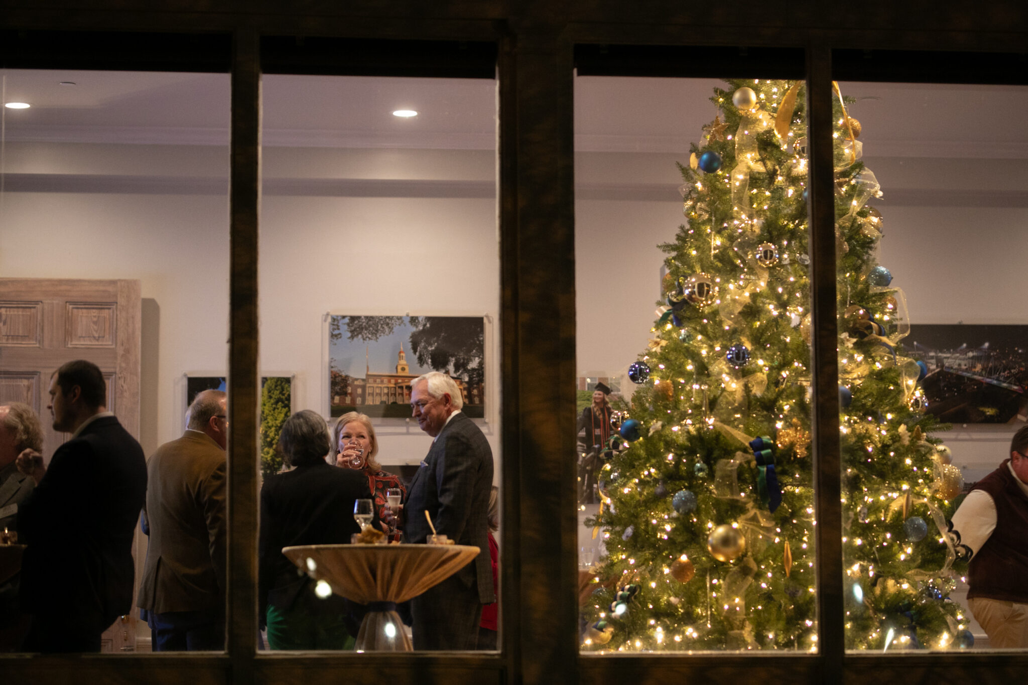 View looking inside a how through large paned windows. Inside are a Christmas tree and people socializing.