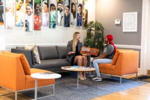 An admissions counselor sits on a gray sofa next to a prospective student in an orange chair showing a Mercer brochure. Pictures of Mercer students hang on the wall behind them.