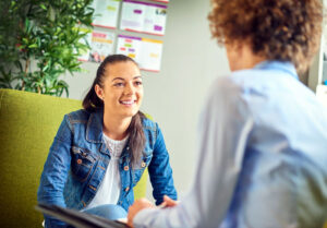 Two individuals having a discussion in an office setting, one facing the camera smiling, surrounded by plants and posters on the wall.
