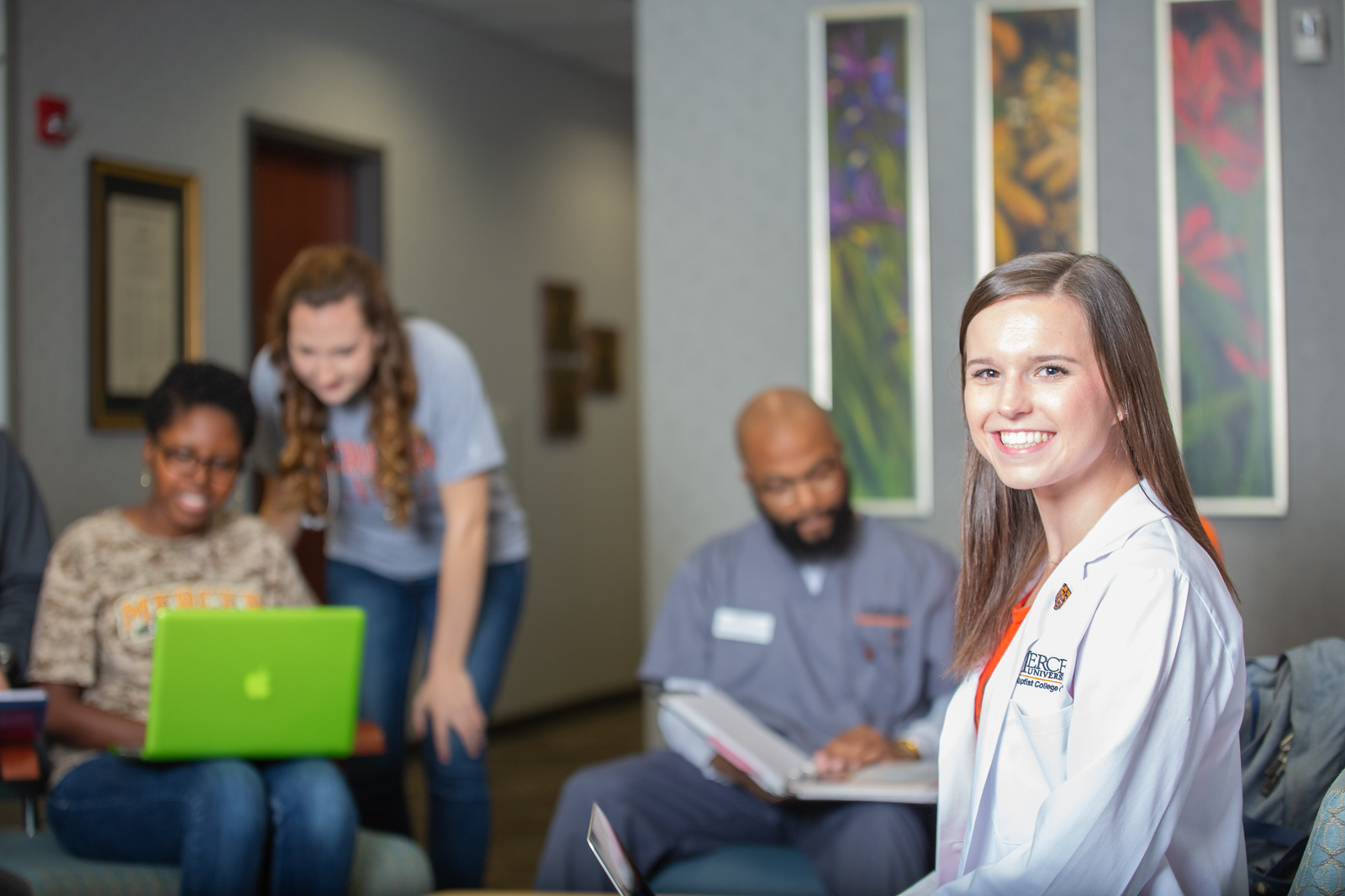 A smiling student looks at the camera and wears a white lab coat with the words Mercer University Georgia Baptist College of Nursing in the corner. Three other students wearing Mercer shirts are seen studying in the background.