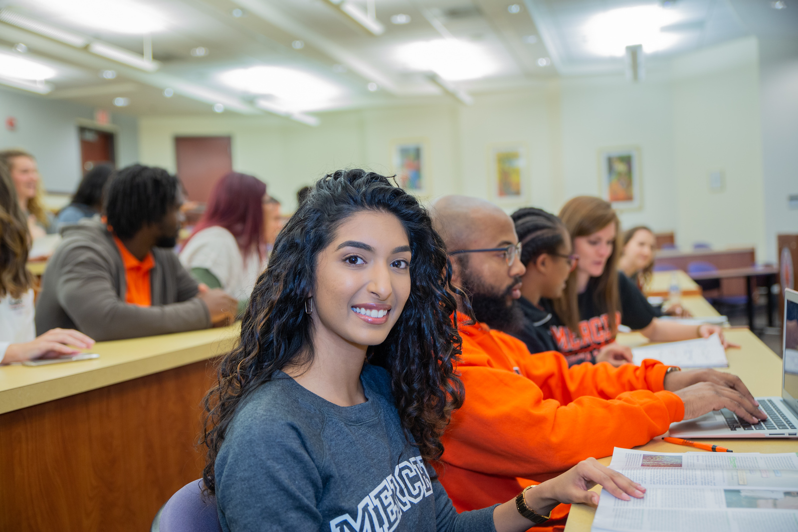 A student wearing a Mercer T-shirt smiles at the camera. She is sitting at a long desk in a lecture hall where other students wearing orange and black are working.