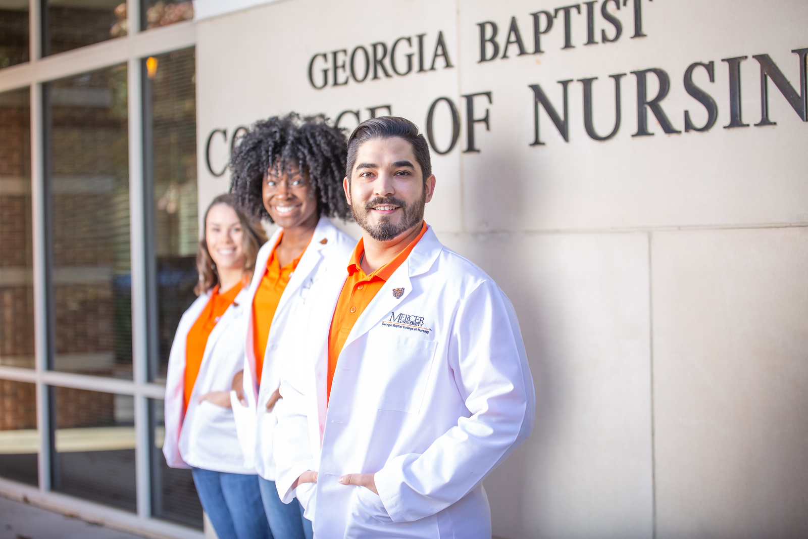 Three students wearing orange shirts under a white coat stand in front of a sign that says, "Georgia Baptist College of Nursing."