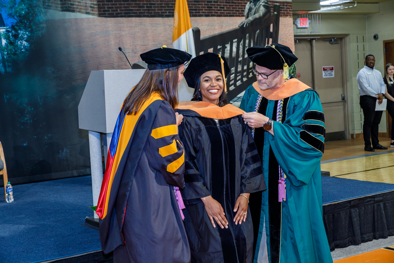 Two people wearing academic regalia place an apricot-colored hood on a graduate during commencement.